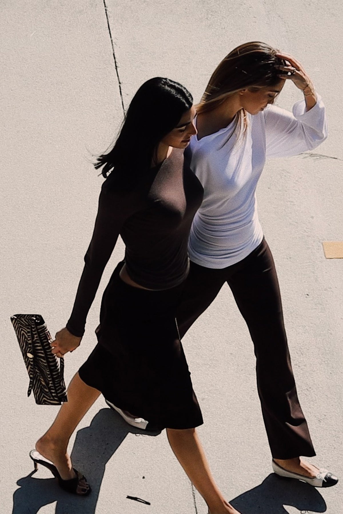 Two women walking outdoors on a sunny day, one wearing a dark brown long-sleeve crop top and black skirt holding a black and white patterned clutch, and the other wearing a white long-sleeve top and brown pants.