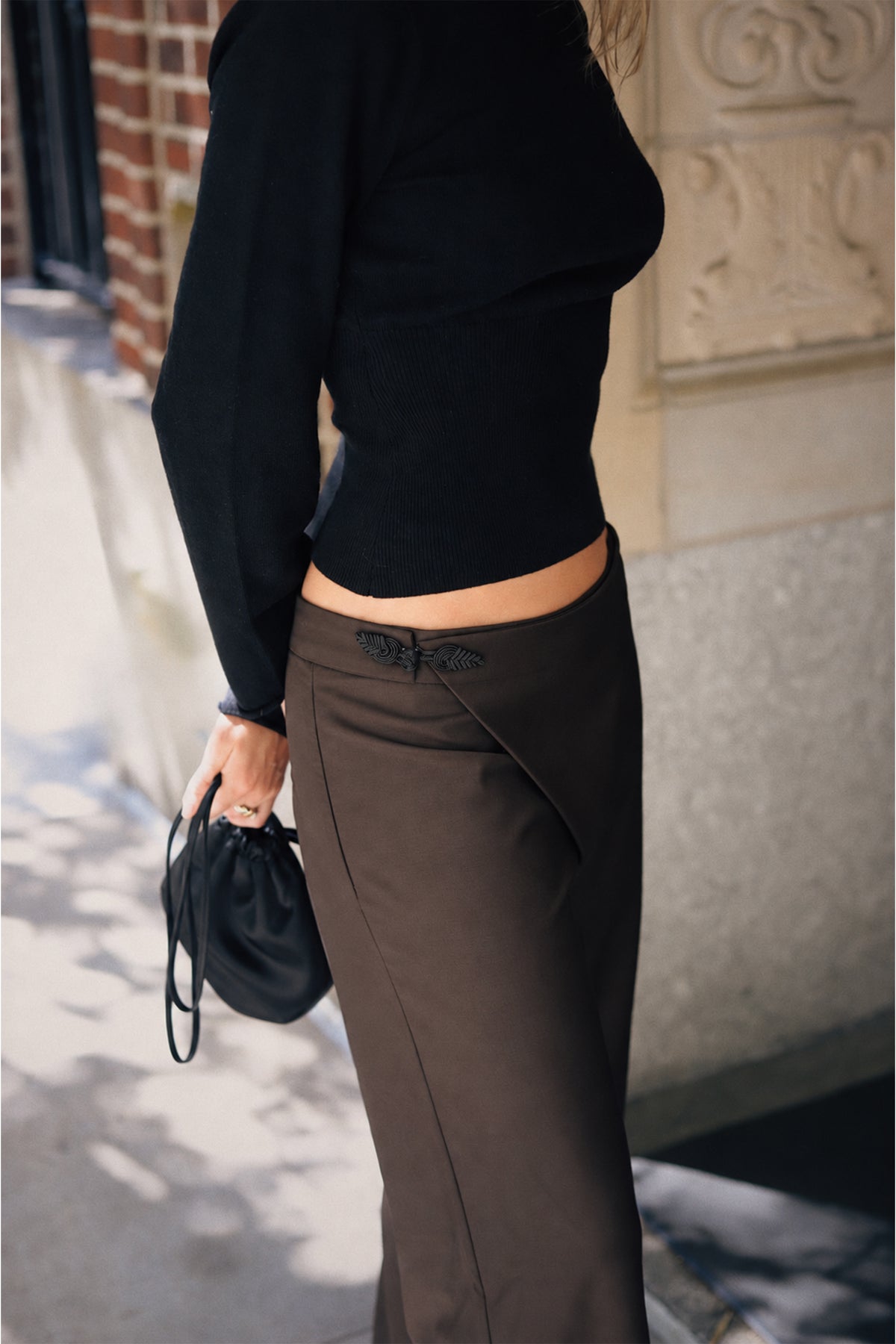 A person wearing a fitted black long-sleeve top and dark brown draped skirt, holding a small black handbag on a city sidewalk.