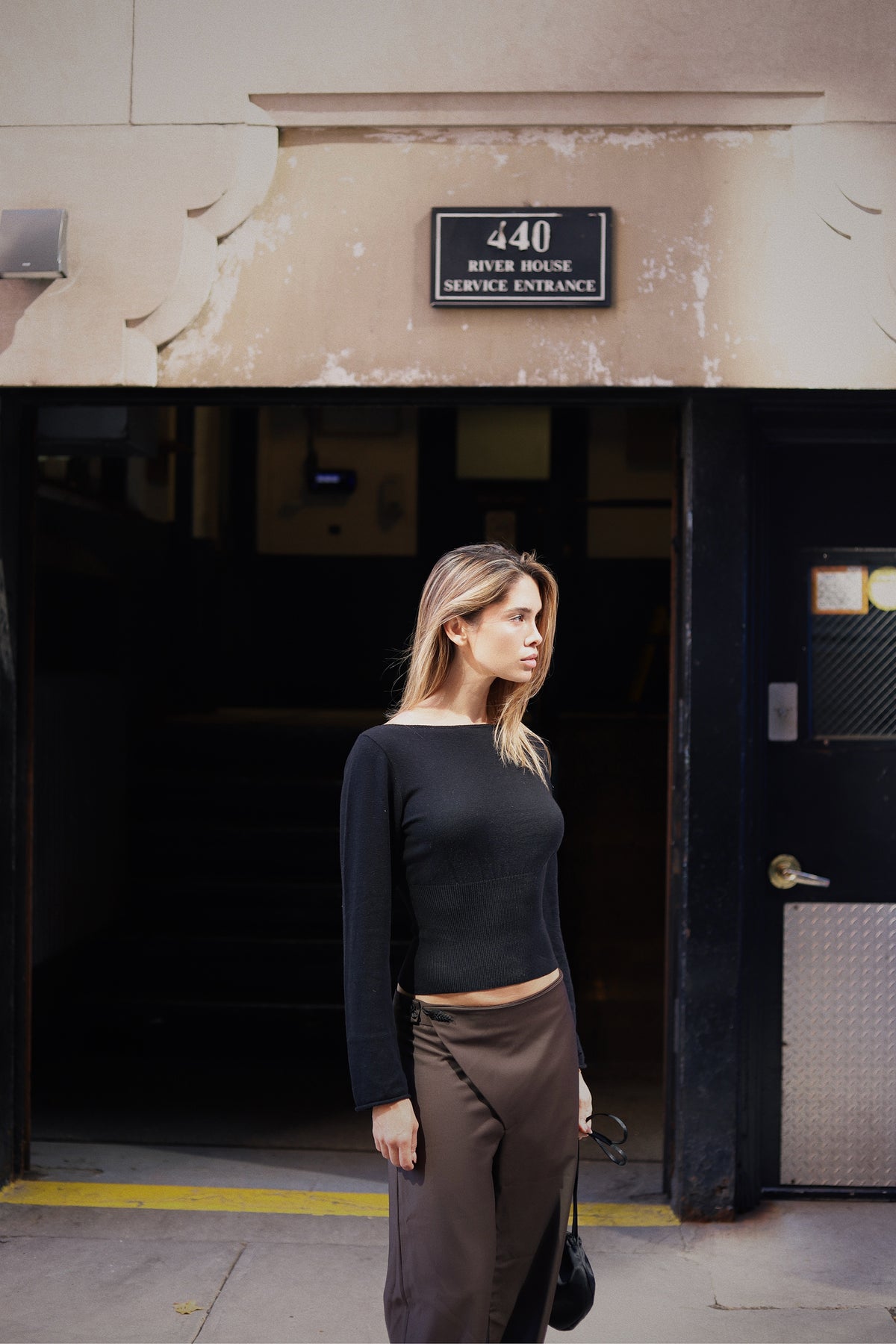 Model stands in front of a building entrance wearing a black long-sleeve knit and brown pants.