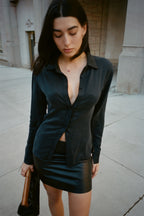 A woman wearing a black satin long-sleeve button-up shirt and a black leather mini skirt, standing on a city sidewalk.