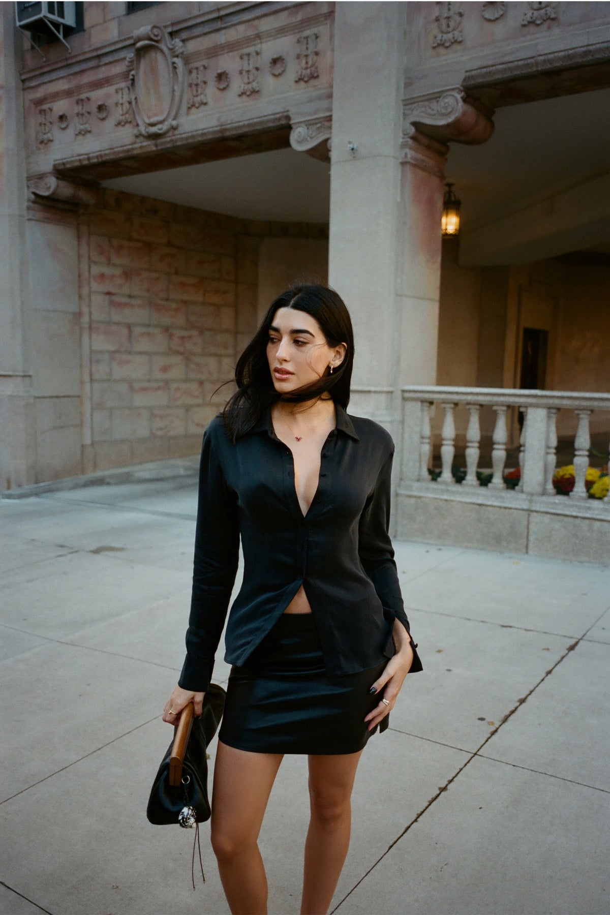 A woman stands outdoors in front of a stone building wearing a black satin long-sleeve collared mini dress with a front slit and a matching black mini skirt. She is holding a black handbag.