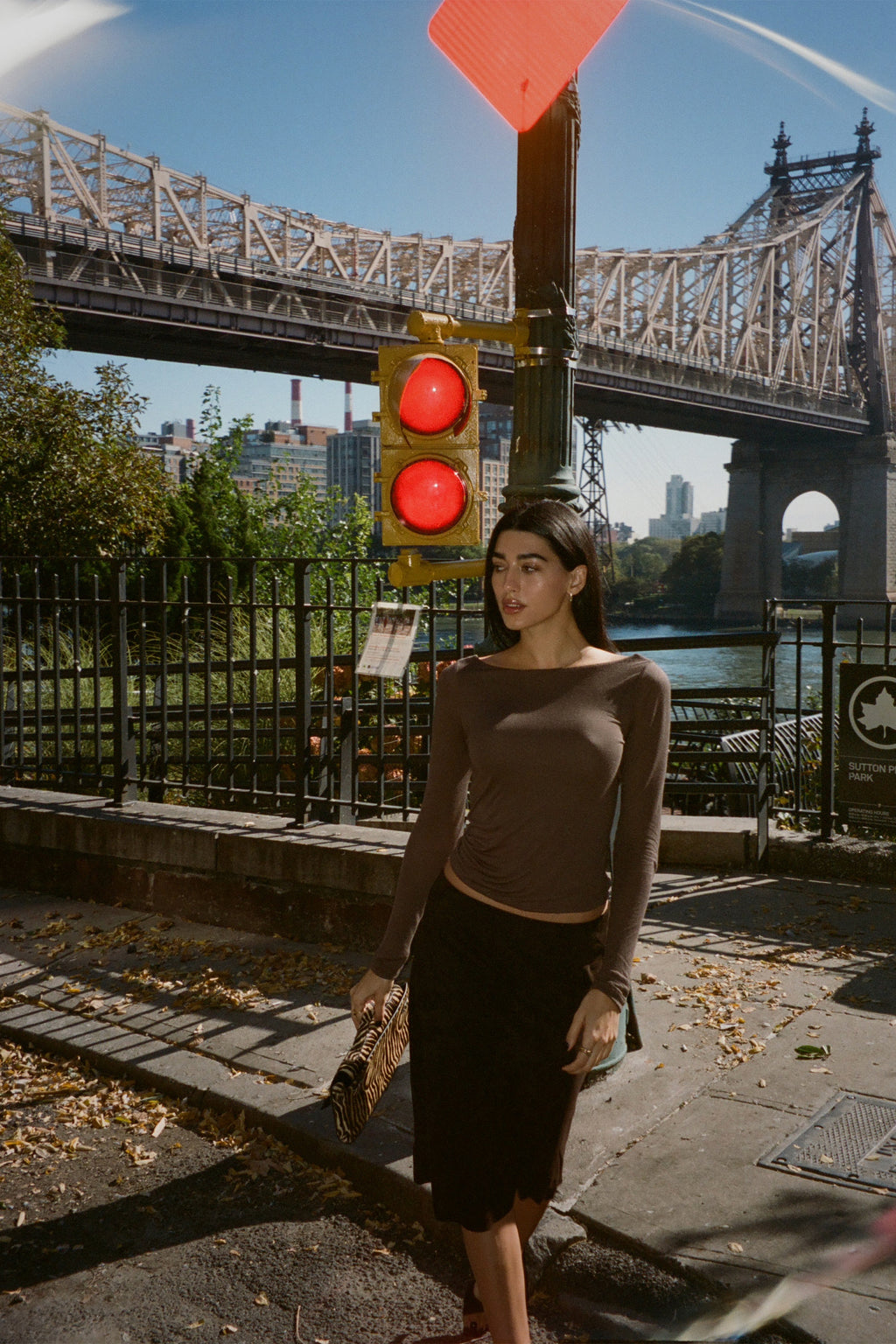 A model in New York City sidewalk in front of a large steel bridge and traffic light, wearing a fitted brown long-sleeve top and a suede midi skirt, holding a small patterned clutch.