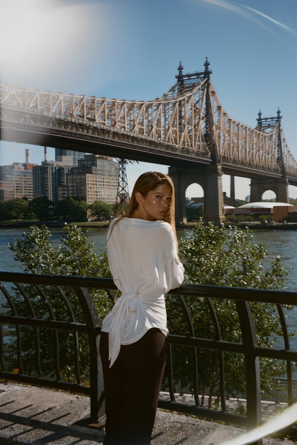 A woman wearing a white long-sleeve wrap-style top and dark pants stands by a waterfront railing with a large bridge and city skyline in the background.