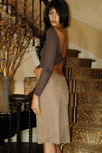 A woman wearing a brown long-sleeve backless crop top and a taupe suede skirt, standing 
