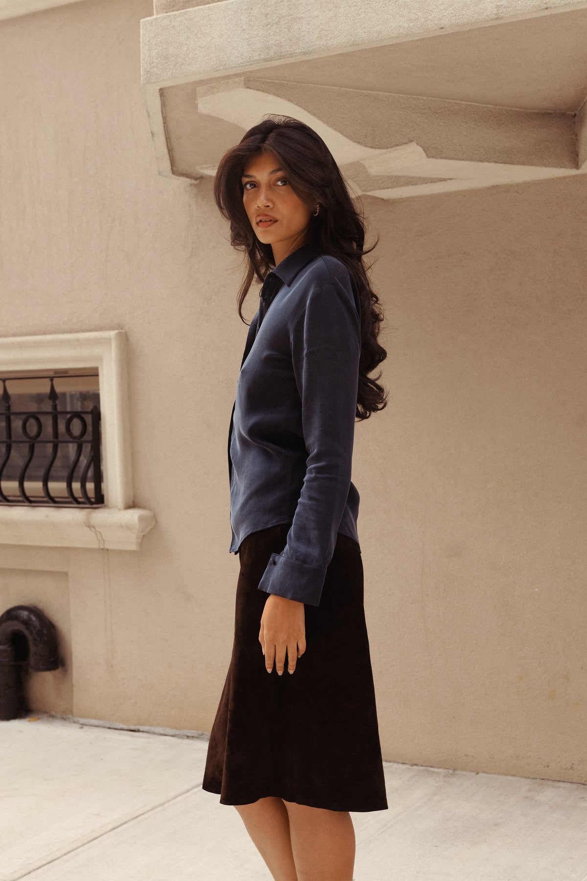 A woman wearing a dark blue long-sleeve button-up shirt and a dark brown suede midi skirt.