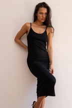 A woman modeling a black midi slip dress against a plain off-white wall.