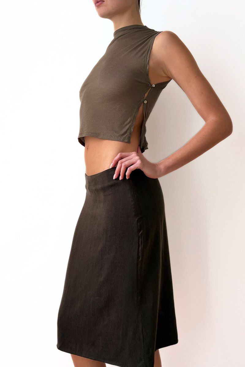 Model wearing a sleeveless olive green top and brown skirt against a white wall.