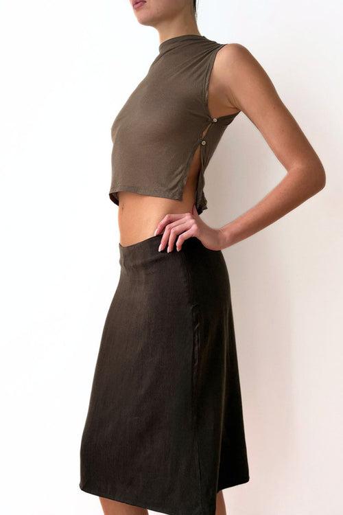 Model wearing a sleeveless olive green top and brown skirt against a white wall.
