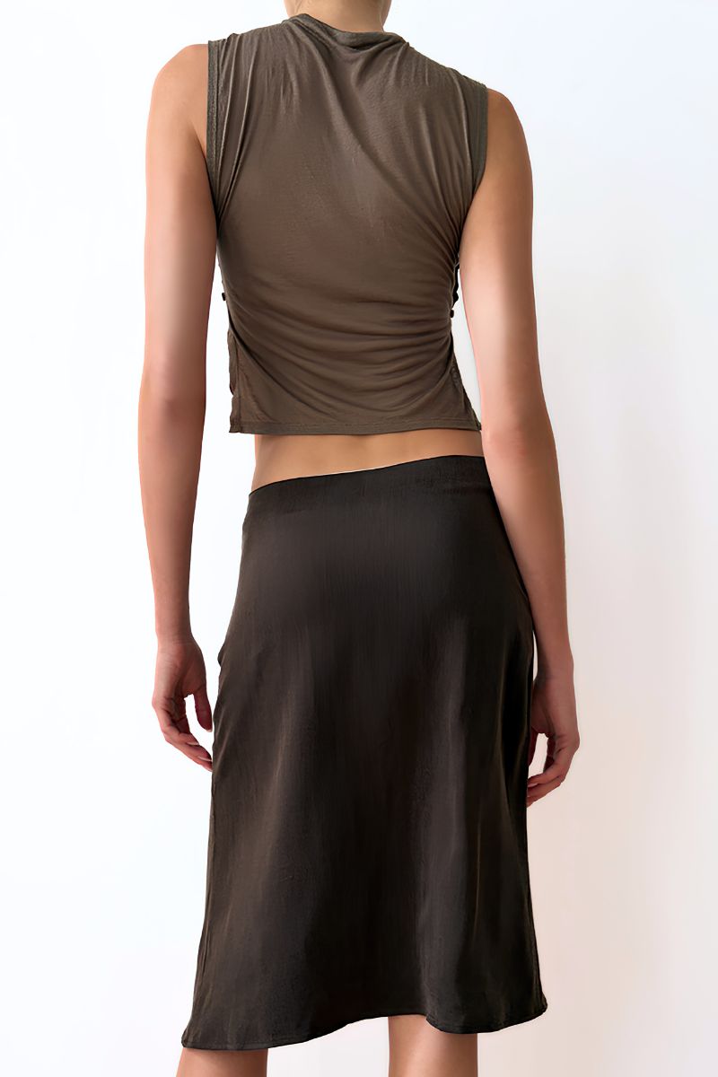 Model wearing a sleeveless olive green top and brown skirt against a white wall.