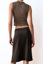 Model wearing a sleeveless olive green top and brown skirt against a white wall.
