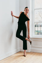 Woman in a black one-shoulder top and pants posing in a room with a window.