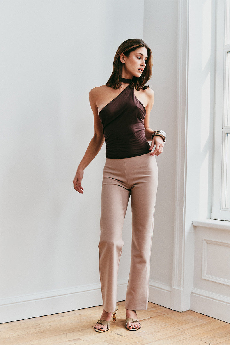 Woman wearing a brown one-shoulder top and beige pants in a room with white walls and wooden flooring.