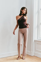 Woman wearing a brown one-shoulder top and beige pants in a room with white walls and wooden flooring.