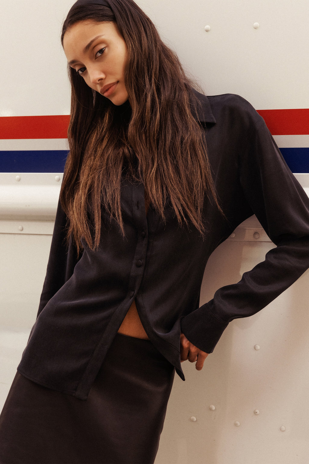 A woman wearing a black satin long-sleeve button-up shirt and a matching black leather skirt, leaning against a white wall with red and blue stripes.