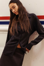 A woman wearing a black satin long-sleeve button-up shirt and a matching black leather skirt, leaning against a white wall with red and blue stripes.