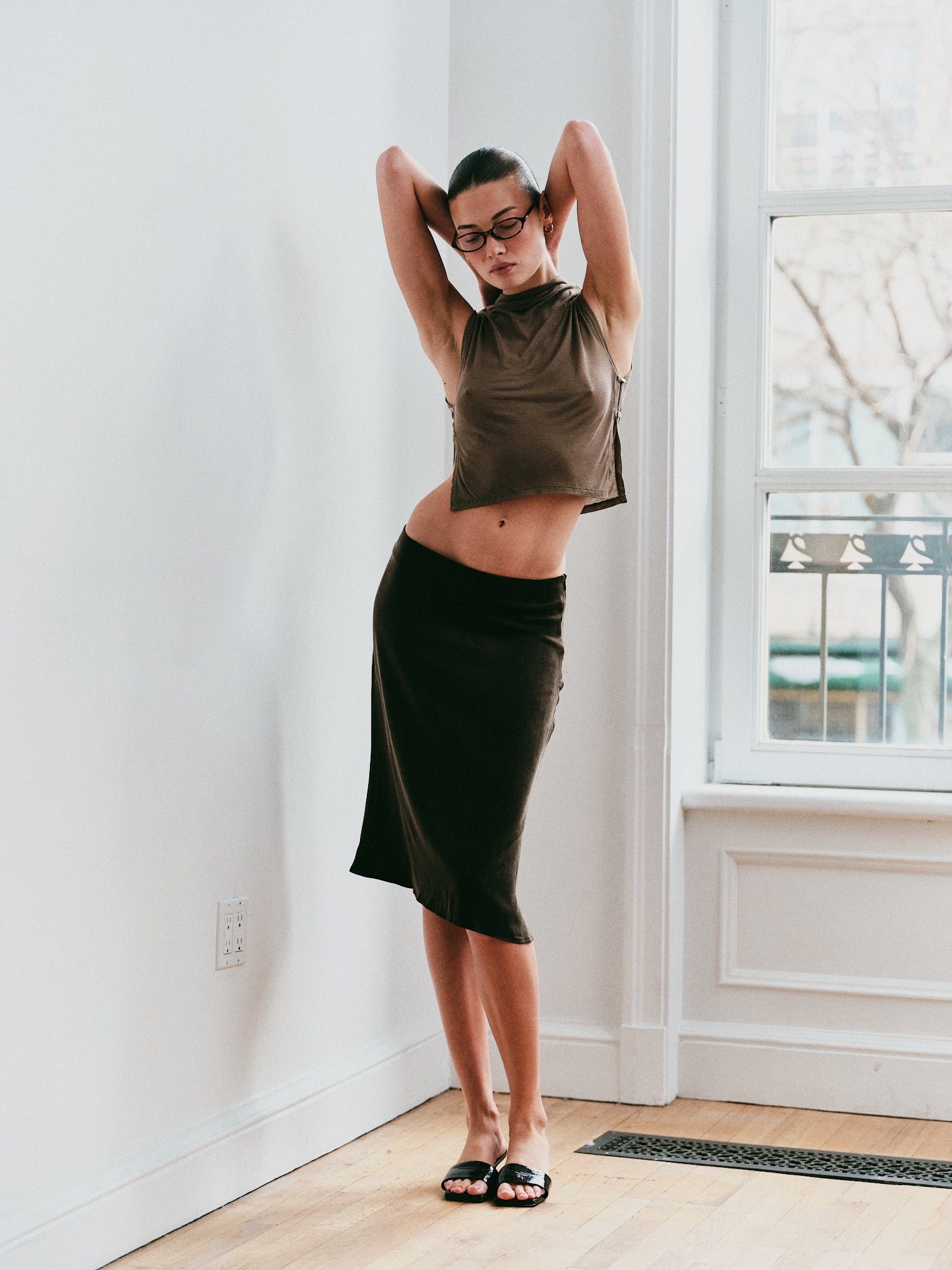 Model wearing a green top and brown skirt standing in a room with a window.