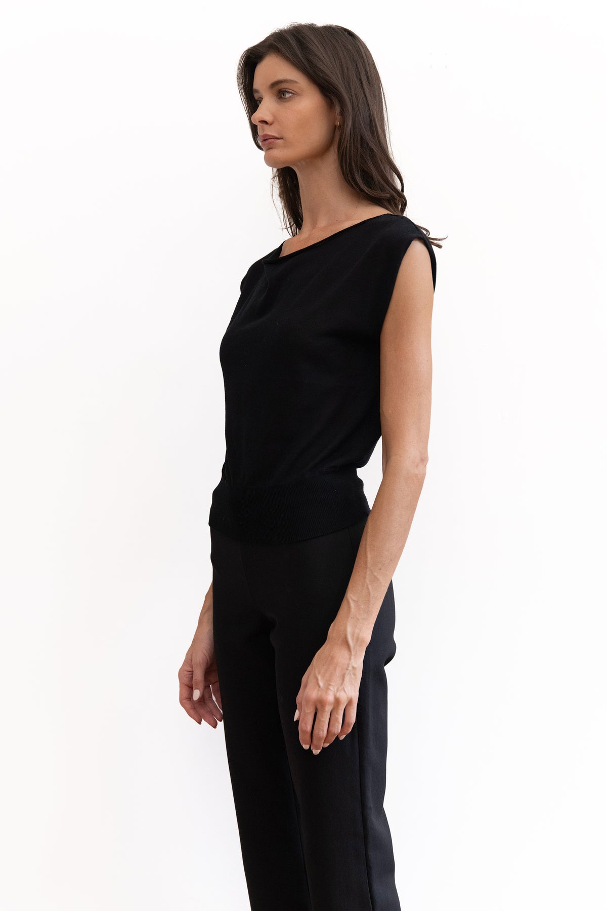 A woman modeling a black sleeveless top and black trousers, shown in profile against a plain white studio background.