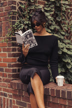 A woman wearing a black off-the-shoulder long-sleeve top and matching skirt sits on a brick ledge next to a brick wall with green ivy, reading a book and holding a white coffee cup.