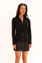 A model wearing a black long-sleeve button-front shirt with a low open neckline and a matching black leather mini skirt, 