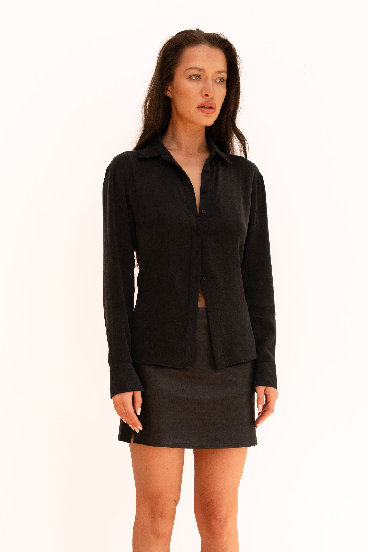 A model wearing a black long-sleeve button-front shirt with a low open neckline and a matching black leather mini skirt, 