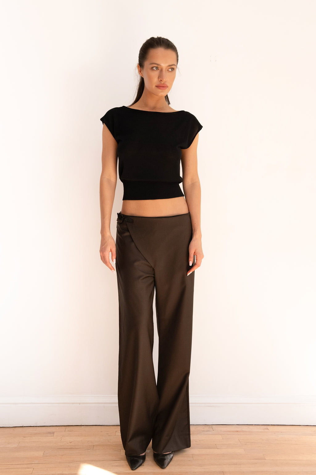 A woman stands against a plain white wall wearing a black cropped short-sleeve top and dark brown wide-leg pants, paired with black pointed shoes.