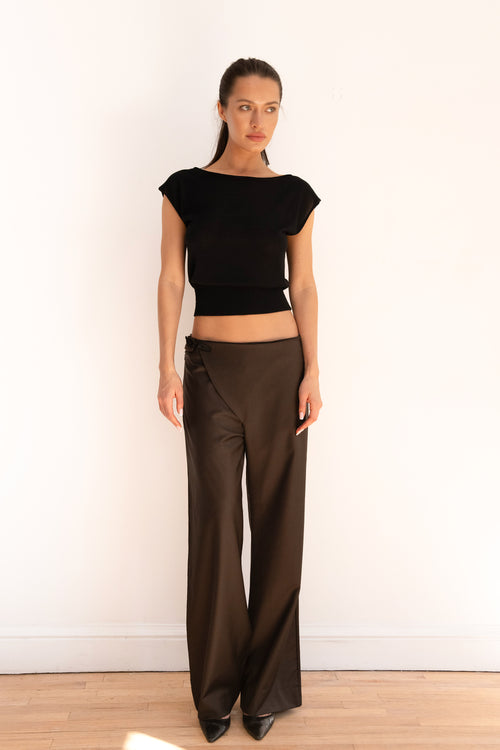 A woman stands against a plain white wall wearing a black cropped short-sleeve top and dark brown wide-leg pants, paired with black pointed shoes.