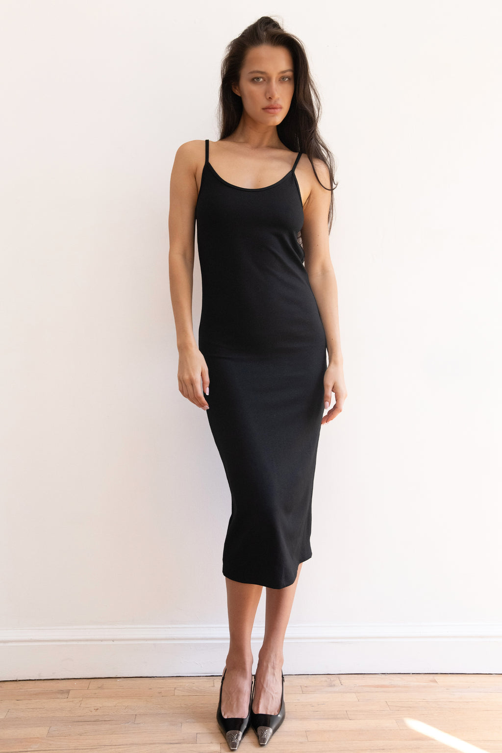 A woman modeling a black midi slip dress with a fitted silhouette, paired with black pointed-toe heels, standing against a plain white wall on a light wood floor.