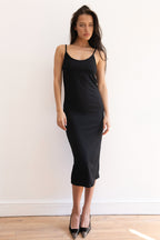 A woman modeling a black midi slip dress with a fitted silhouette, paired with black pointed-toe heels, standing against a plain white wall on a light wood floor.
