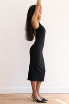 A woman modeling a black midi slip dress with a fitted silhouette, paired with black pointed-toe heels, standing against a plain white wall on a light wood floor.