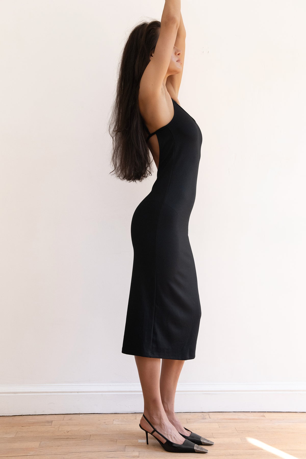 A woman modeling a black midi slip dress with a fitted silhouette, paired with black pointed-toe heels, standing against a plain white wall on a light wood floor.