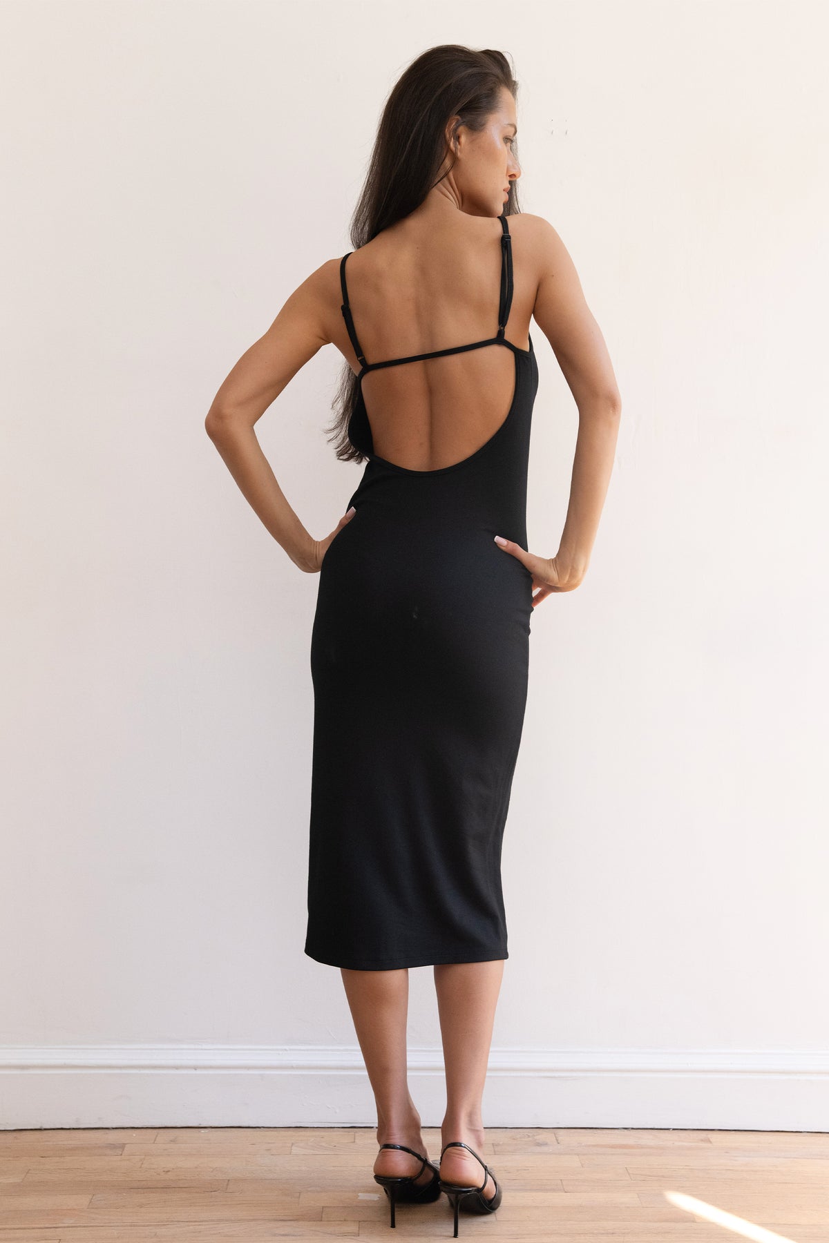 A woman modeling a black midi slip dress with a fitted silhouette, paired with black pointed-toe heels, standing against a plain white wall on a light wood floor.