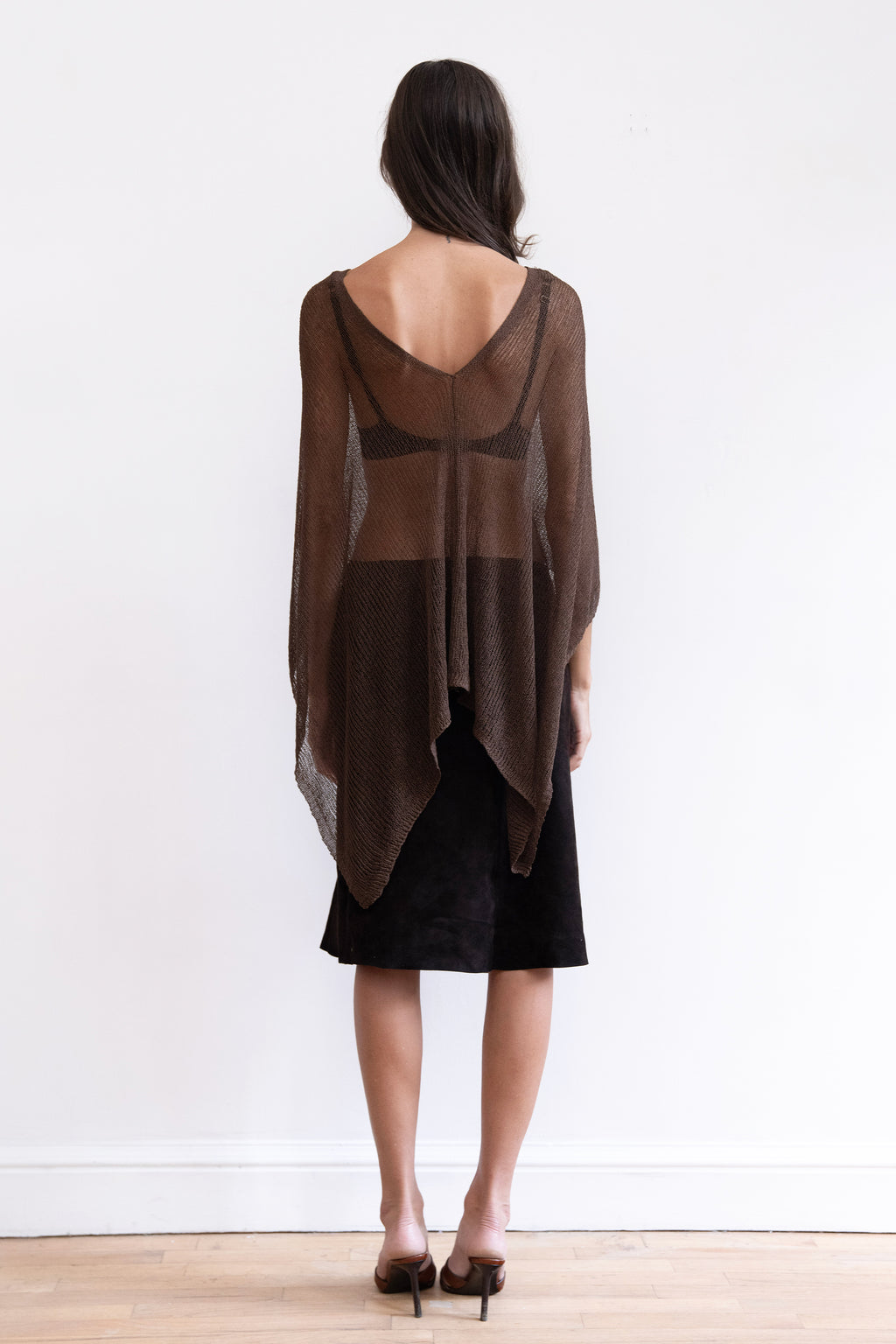 A model wearing a sheer dark brown long-sleeve shawl knit top with a black midi skirt and brown heeled sandals.
