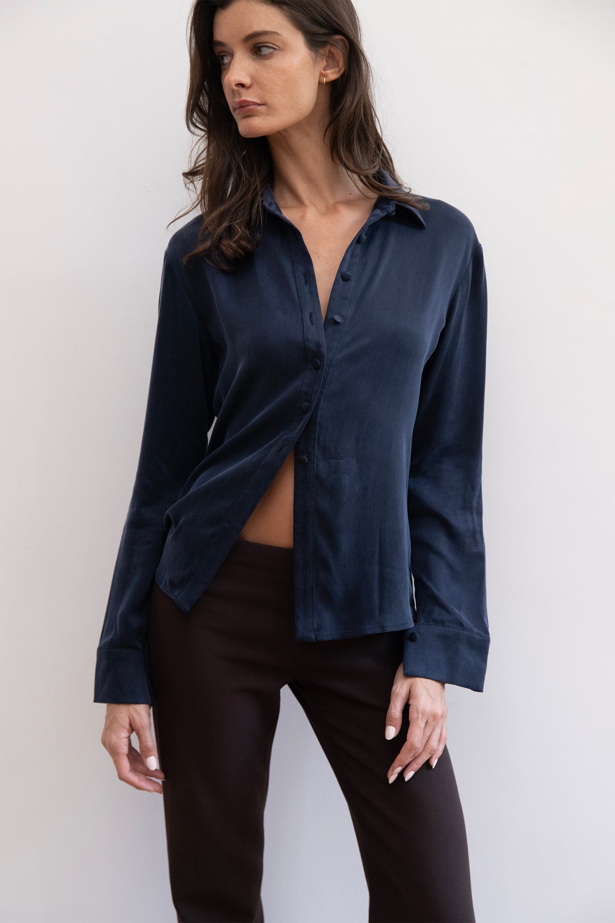 A model wearing a dark navy blue satin button-up shirt with a twisted front detail, paired with dark brown pants, against a plain light gray background.