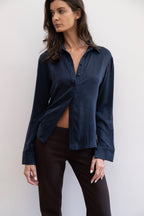 A woman modeling a dark navy blue satin button-up shirt with a twisted front detail, paired with dark brown pants, against a plain light gray background.
