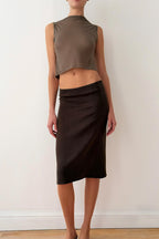 Model wearing a sleeveless olive green top and brown skirt against a white wall.