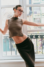 Model in a green tank top and black pants standing in front of a window with a blurred outdoor view.